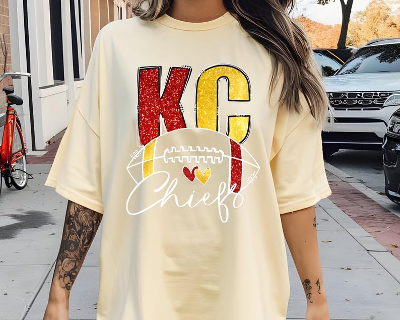 May include: Cream-colored t-shirt featuring the text "KC Chiefs" in red and yellow. The design includes a football graphic with red and yellow hearts. The shirt has short sleeves and a relaxed fit.