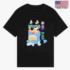 May include: Black t-shirt featuring a cartoon dog character in blue, yellow, and black with the text "GIRL DAD" in colorful letters. The shirt also has a small "MADE IN USA" label with the American flag.