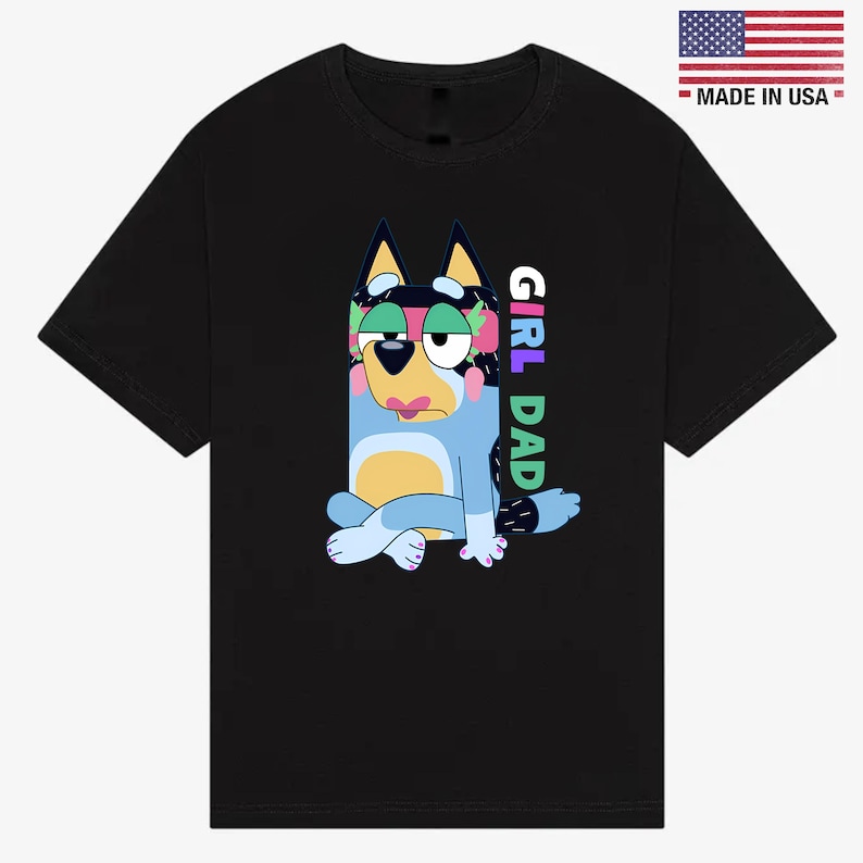May include: Black t-shirt featuring a cartoon dog character in blue, yellow, and black with the text "GIRL DAD" in colorful letters. The shirt also has a small "MADE IN USA" label with the American flag.