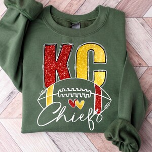 May include: Green sweatshirt with the letters "KC" in red glitter and yellow, with a football graphic and the word "Chiefs" in white script. The football has red and yellow accents and two heart shapes.