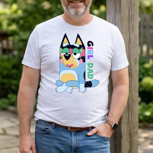 May include: White t-shirt featuring a cartoon dog character in blue, black, and yellow with a floral pattern on its head. The text "GIRL DAD" is printed vertically on the right side of the shirt in colorful letters. The shirt is worn by a person.