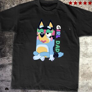 May include: Black t-shirt featuring a cartoon dog character in blue, yellow, and pink. The dog is sitting with its legs crossed and tongue sticking out. The words "GIRL DAD" are printed vertically on the right side of the shirt.