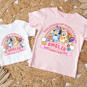 May include: Two children's t-shirts, one white and one pink, featuring a colorful graphic. The graphic includes the text "THIS EPISODE OF BLUEY IS CALLED AMELIA TURNS TWO!" with cartoon characters, a rainbow, cake, and balloons.