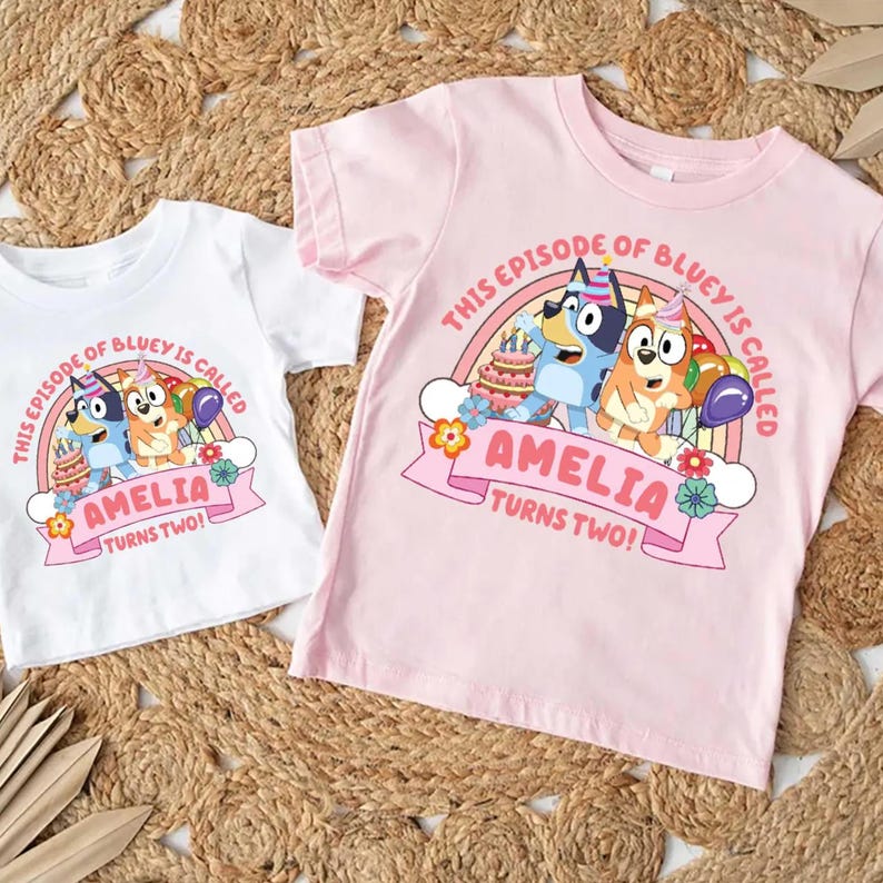May include: Two children's t-shirts, one white and one pink, featuring a colorful graphic. The graphic includes the text "THIS EPISODE OF BLUEY IS CALLED AMELIA TURNS TWO!" with cartoon characters, a rainbow, cake, and balloons.