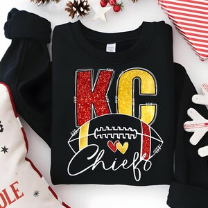 May include: Black crewneck sweatshirt with a graphic design. The design features the letters "KC" in red glitter and gold, a football, and the word "Chiefs" in white script. Two hearts are also included in the design.