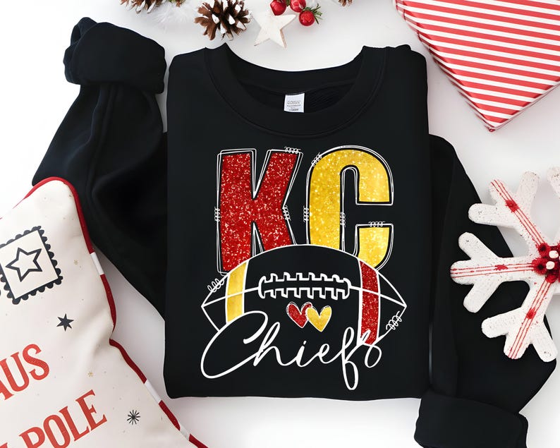 May include: Black crewneck sweatshirt with a graphic design. The design features the letters "KC" in red glitter and gold, a football, and the word "Chiefs" in white script. Two hearts are also included in the design.