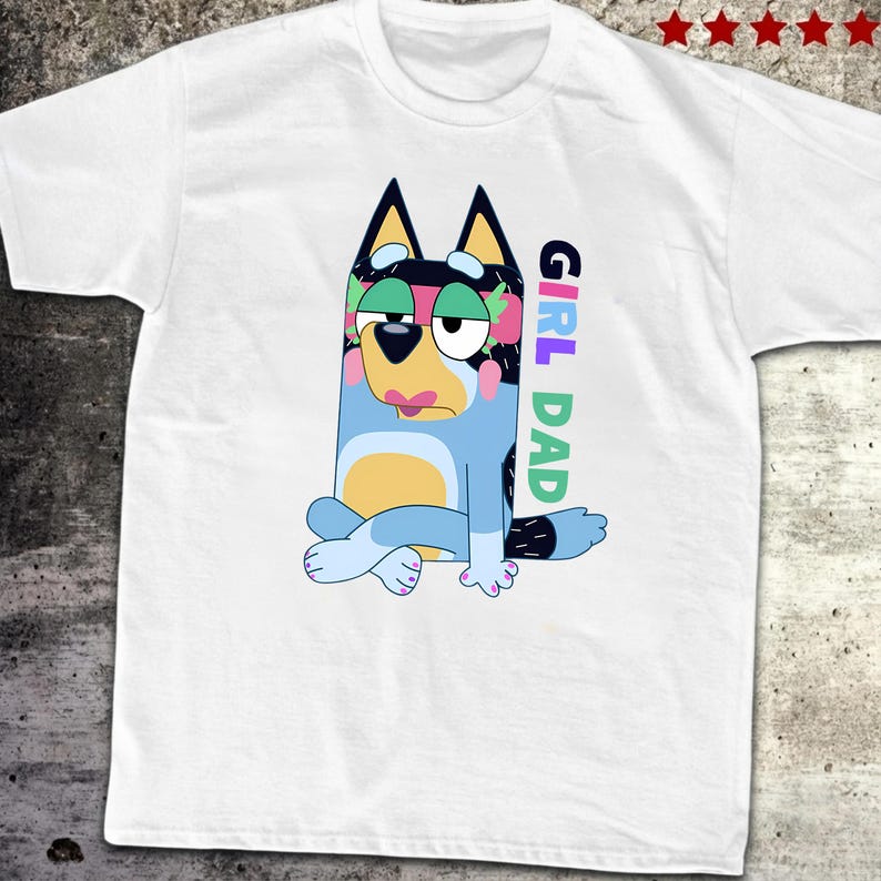 May include: White t-shirt featuring a cartoon dog character in blue, black, and yellow. The dog is sitting with its legs crossed and has a sad expression. The words "GIRL DAD" are printed vertically on the right side of the shirt in colorful letters.