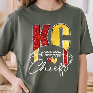May include: Olive green t-shirt featuring the letters "KC" in red and yellow glitter, with a football graphic and the word "Chiefs" in white script. Two hearts are in the center of the football graphic.