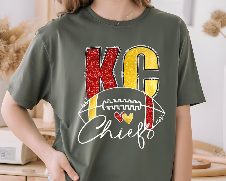 May include: Olive green t-shirt featuring the letters "KC" in red and yellow glitter, with a football graphic and the word "Chiefs" in white script. Two hearts are in the center of the football graphic.