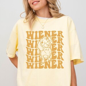 K&ouml;nnte beinhalten: Ein gelbes T-Shirt mit einer Grafik im Retro-Stil eines Dackelhundes und dem Text "WIENER", der in einem gestapelten Muster wiederholt wird.