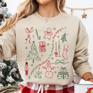 May include: A sand-colored sweatshirt with a festive Christmas design. The design includes red and green illustrations of a Christmas tree, snowman, gifts, and the text "all I want for Christmas is my students to stop saying 6-7".