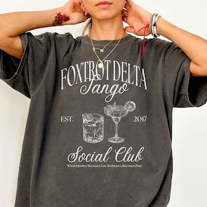 Op de afbeelding: Een donkergrijs T-shirt met een wit grafisch ontwerp. De grafiek toont de tekst "Fox Trot Delta Sango" met de woorden "Social Club" eronder. De grafiek bevat ook twee illustraties van cocktails, een met een limoenwig en de andere met ijsblokjes. De tekst "Est. 2017" is boven de cocktails gedrukt. De tekst "When Injustice Becomes Law, Resistance Becomes Duty" is onder de cocktails gedrukt.