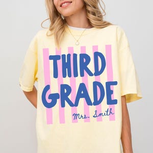 May include: A light yellow t-shirt with the words "THIRD GRADE" in blue, and pink vertical stripes. The name "Mrs. Smith" is below the words. The word "BANANA" is in a yellow box in the bottom left corner.