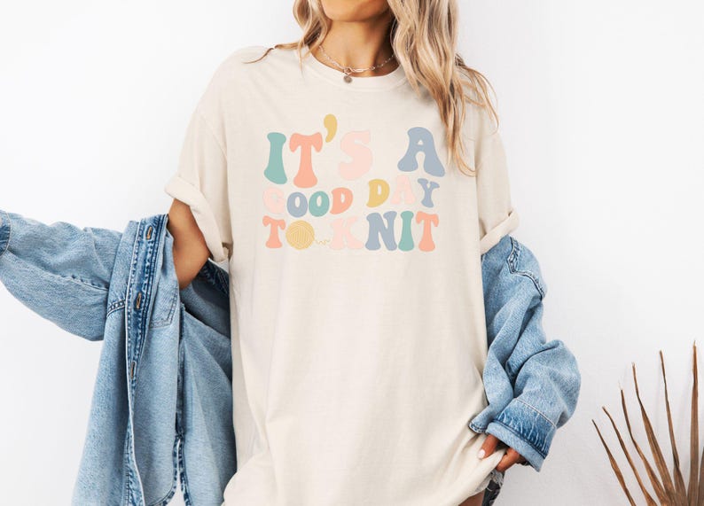 May include: A white t-shirt with the text "It's a good day to knit" in colorful, retro-style lettering.