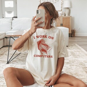 Puede incluir: Una camiseta blanca con un gr&aacute;fico rojo de un gato sentado en una computadora port&aacute;til. El texto "I work on computers" est&aacute; impreso en rojo debajo de la imagen.