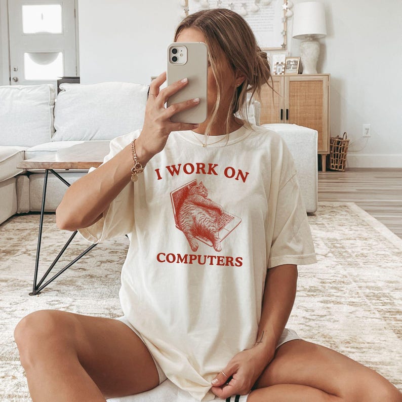 Puede incluir: Una camiseta blanca con un gr&aacute;fico rojo de un gato sentado en una computadora port&aacute;til. El texto "I work on computers" est&aacute; impreso en rojo debajo de la imagen.
