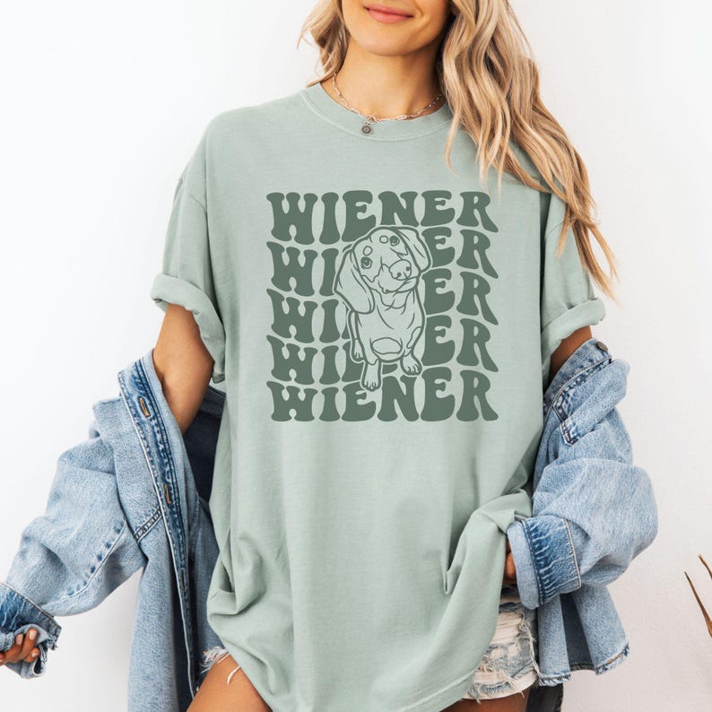 K&ouml;nnte beinhalten: Ein salbeigr&uuml;nes T-Shirt mit einer Grafik eines Dackels und dem Text "Wiener", der in einer Retro-Schrift wiederholt wird.
