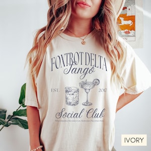 Op de afbeelding: Ivoorwit T-shirt met een grafisch ontwerp met de tekst "Fox Trot Delta Tango Social Club" met de jaren "EST. 2017" en twee illustraties van cocktails. De tekst "When Injustice Becomes Law, Resistance Becomes Duty" is onder de afbeelding gedrukt.