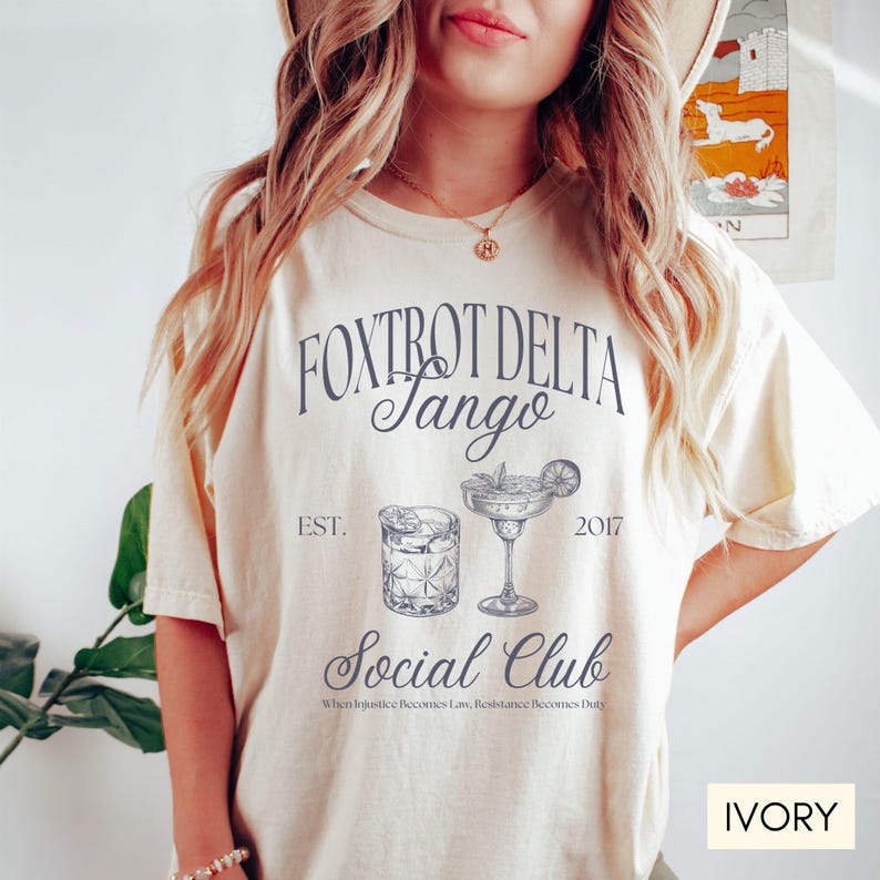 Op de afbeelding: Ivoorwit T-shirt met een grafisch ontwerp met de tekst "Fox Trot Delta Tango Social Club" met de jaren "EST. 2017" en twee illustraties van cocktails. De tekst "When Injustice Becomes Law, Resistance Becomes Duty" is onder de afbeelding gedrukt.