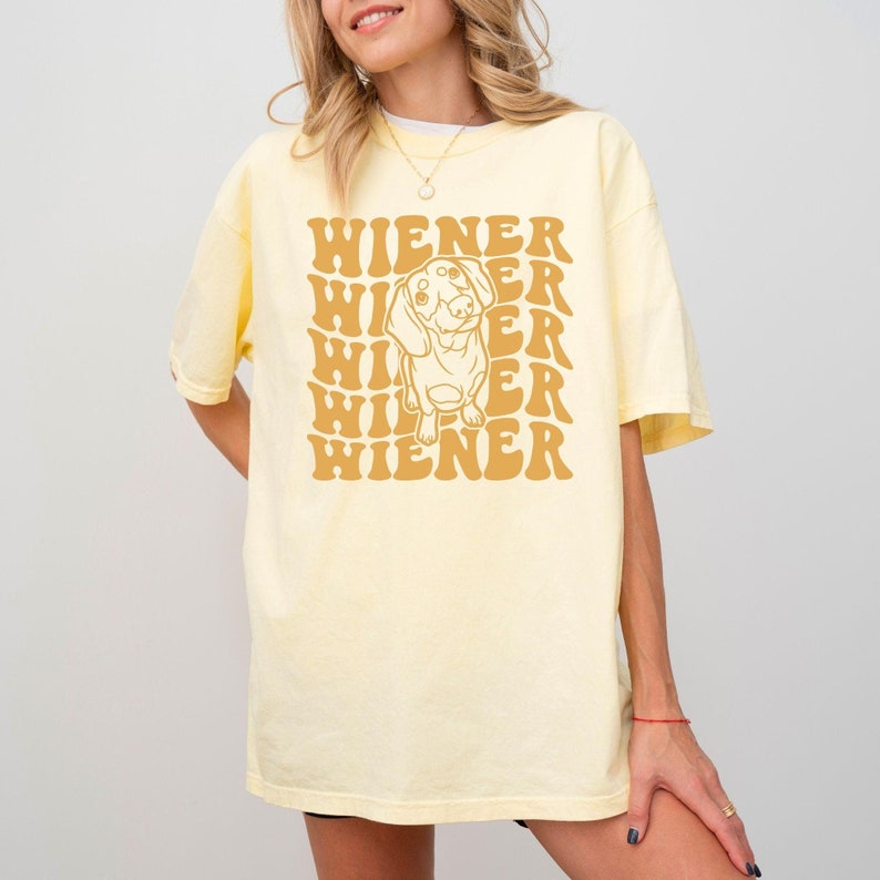K&ouml;nnte beinhalten: Ein gelbes T-Shirt mit einer Grafik im Retro-Stil eines Dackelhundes und dem Text "WIENER", der in einem gestapelten Muster wiederholt wird.