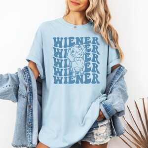 K&ouml;nnte beinhalten: Ein hellblaues T-Shirt mit einer Grafik eines Dackelhundes und dem Text "WIENER", der in einer Retro-Schrift wiederholt wird.