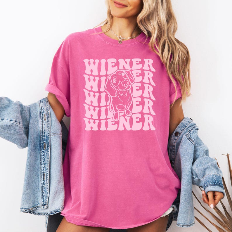 K&ouml;nnte beinhalten: Ein pinkfarbenes T-Shirt mit einer wei&szlig;en Umrandung eines Dackels und dem Text "WIENER", der in einer Retro-Schrift wiederholt wird.