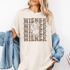 K&ouml;nnte beinhalten: Ein beiges T-Shirt mit einer Grafik eines Dackelhundes und dem Text "WIENER", der in einer Retro-Schrift wiederholt wird.