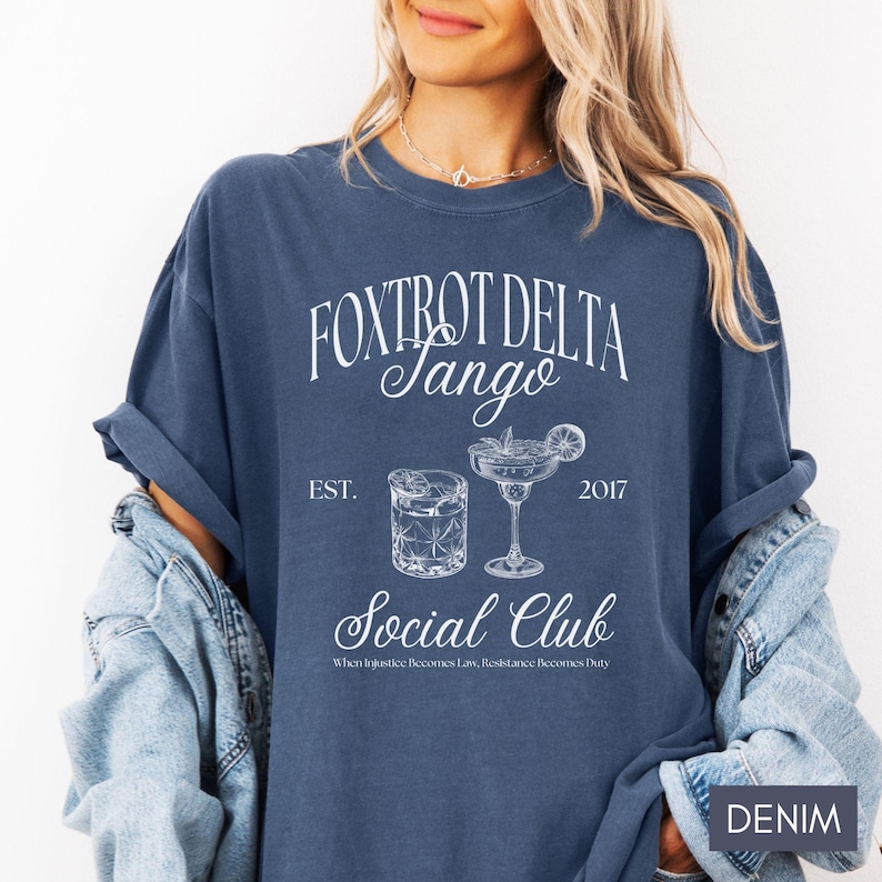 Op de afbeelding: Een blauw T-shirt met witte tekst die "Fox Trot Delta Jango Est. 2017 Social Club When Injustice Becomes Law, Resistance Becomes Duty" luidt. Het shirt heeft twee illustraties van cocktails, een met een limoenwig en de andere met een kers.