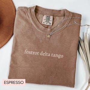 May include: A brown t-shirt with the text "foxtrot delta tango" printed in white. The shirt is labeled "Espresso" and has a tag that says "Comfort Colors".