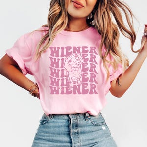 K&ouml;nnte beinhalten: Pinkes T-Shirt mit einem Grafikdruck eines Dackelhundes und dem Text "WIENER", der in einer Retro-Schrift wiederholt wird.