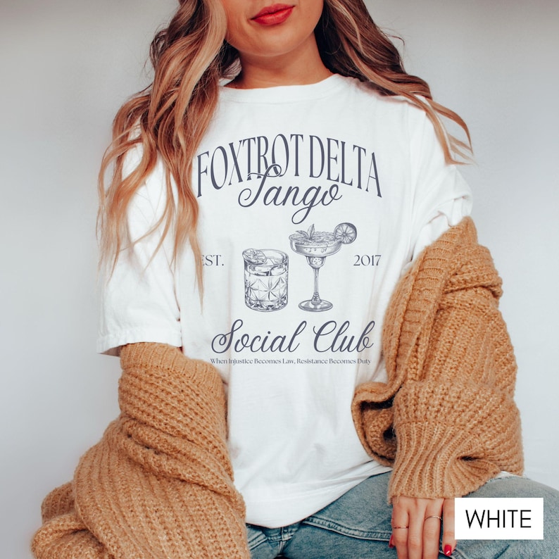 Op de afbeelding: Wit T-shirt met een grafisch ontwerp van twee cocktails en de tekst "FOXTROT DELTA Tango Social Club Est. 2017 When Injustice Becomes Law, Resistance Becomes Duty".