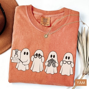 May include: A terracotta-colored t-shirt featuring a graphic of four white ghosts. One ghost holds an eye chart, another wears glasses, the third holds binoculars, and the fourth wears spectacles. The shirt has a Comfort Colors brand label at the neck.