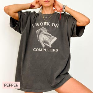 Puede incluir: Una camiseta gris oscura de gran tama&ntilde;o con el texto "Trabajo en computadoras" y un gato de dibujos animados sentado en una computadora port&aacute;til.