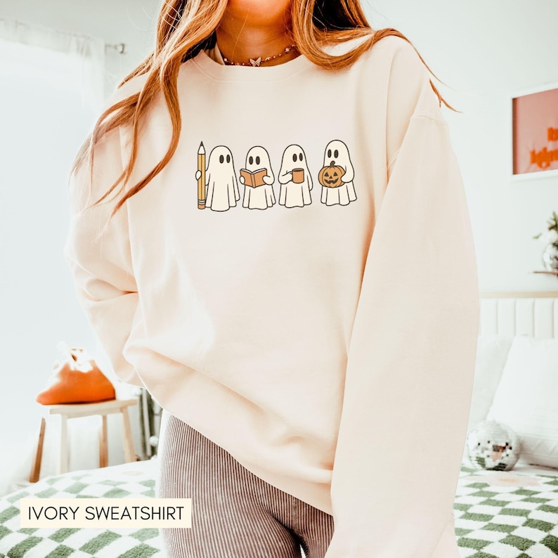 May include: Ivory sweatshirt featuring a line of cartoon ghosts, each holding a pencil, book, coffee cup, and pumpkin. The design is in a minimalist style with black outlines and a retro aesthetic. The text "IVORY SWEATSHIRT" is at the bottom.