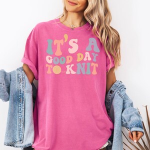 May include: A pink t-shirt with the text "It's a good day to knit" in a retro font with colorful letters.