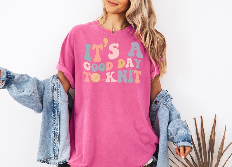 May include: A pink t-shirt with the text "It's a good day to knit" in a retro font with colorful letters.