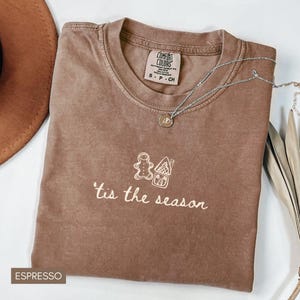 May include: Espresso-colored t-shirt with the phrase "'tis the season" and a gingerbread man and house design. A silver necklace with a pendant rests on the shirt. The shirt is folded and displayed with a brown hat and dried leaves.
