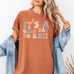 May include: A rust-colored t-shirt with the text "It's a good day to knit" in colorful, retro-style lettering.