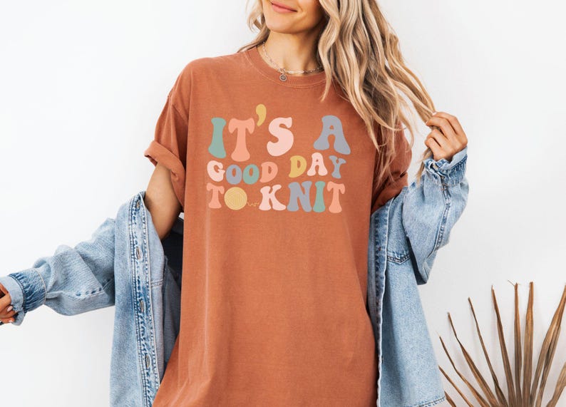 May include: A rust-colored t-shirt with the text "It's a good day to knit" in colorful, retro-style lettering.