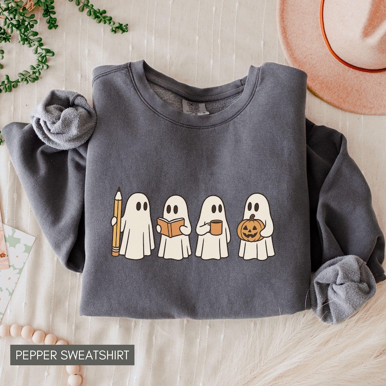 May include: A dark gray sweatshirt with a graphic of four cartoon ghosts, each holding a different item: a pencil, a book, a mug, and a jack-o'-lantern. The text "PEPPER SWEATSHIRT" is at the bottom. The sweatshirt is folded on a light-colored surface.