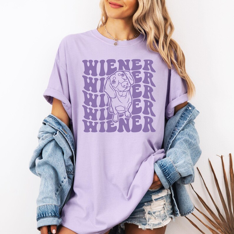 K&ouml;nnte beinhalten: Ein lavendelfarbenes T-Shirt mit einer Grafik eines Dackelhundes und dem Text "WIENER", der in einer Retro-Schrift wiederholt wird.