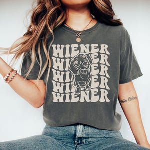 K&ouml;nnte beinhalten: Ein dunkelgraues T-Shirt mit einer Retro-Grafik eines Dackelhundes und dem Text "Wiener", der in einem wellenf&ouml;rmigen Muster wiederholt wird.