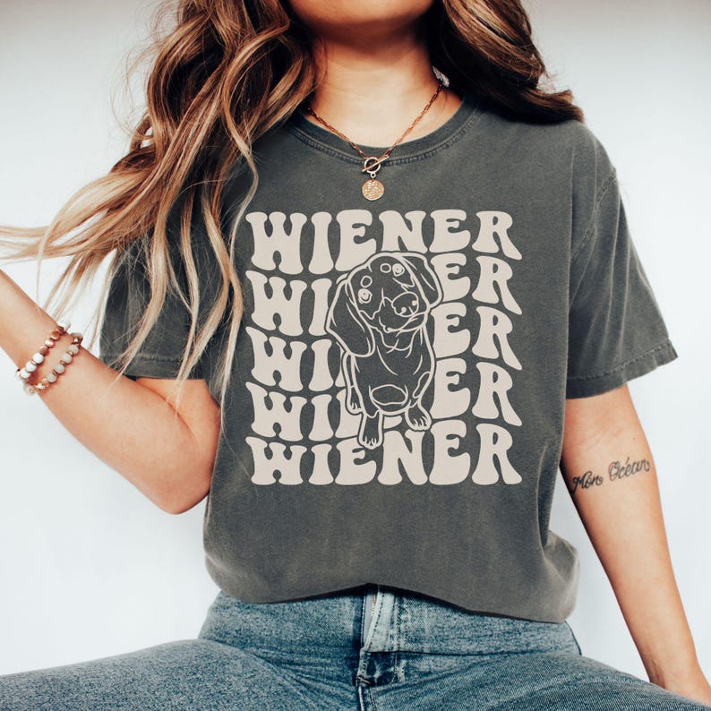 K&ouml;nnte beinhalten: Ein dunkelgraues T-Shirt mit einer Retro-Grafik eines Dackelhundes und dem Text "Wiener", der in einem wellenf&ouml;rmigen Muster wiederholt wird.