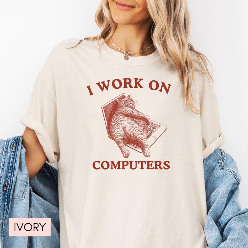 Puede incluir: Camiseta de color marfil con un gr&aacute;fico rojo de un gato sentado en un port&aacute;til. El texto "I work on computers" est&aacute; impreso debajo del gato.