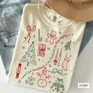 May include: Ivory-colored t-shirt with a Christmas-themed design. The graphic includes a Christmas tree, gingerbread man, snowman, and the text "all I want for christmas is my students to stop saying 6-7".