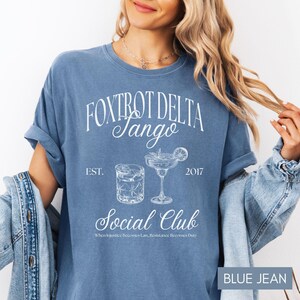Op de afbeelding: Een blauw jeans T-shirt met witte tekst die "Fox Trot Delta Jango Est. 2017 Social Club When Injustice Becomes Law, Resistance Becomes Duty" luidt. Het shirt heeft ook een afbeelding van twee cocktailglazen.