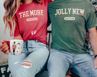 May include: A couple wearing matching Christmas themed t-shirts. The woman's shirt says "The More The Merrier" with a string of lights and baby footprints. The man's shirt says "Jolly New Dad-To-Be" with a string of lights.