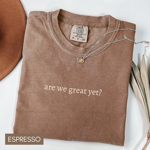 May include: Espresso brown t-shirt with the text "are we great yet?" printed in white. The shirt is made of a soft, comfortable material.