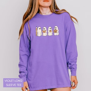 May include: A violet long-sleeve tee featuring a line of cartoon ghosts, each holding a pencil, book, coffee cup, and jack-o'-lantern. The shirt has the text "VIOLET LONG SLEEVE TEE" at the bottom. The shirt is a solid violet color.