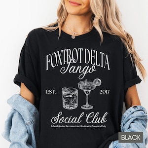 Op de afbeelding: Zwart T-shirt met witte tekst die "FOXTROT DELTA Jango EST. 2017 Social Club When Injustice Becomes Law, Resistance Becomes Duty" luidt. Het shirt heeft twee illustraties van cocktails.